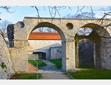 Steinernes Bauwerk mit mehreren Rundbögen, das zu einer historischen Befestigungsanlage gehört. Im Hintergrund sind ein Gebäude mit rotem Ziegeldach und ein schmaler Weg erkennbar, umgeben von Bäumen und Grasflächen.