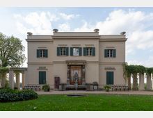 Klassizistisches Gebäude mit weißer Fassade, grünen Fensterläden und einem zentralen Brunnen mit einer Statue in der Mitte. Links und rechts des Gebäudes sind Säulengänge mit bewachsener Pergola sichtbar; im Vordergrund befindet sich eine Rasenfläche mit einem kleinen runden Brunnen.