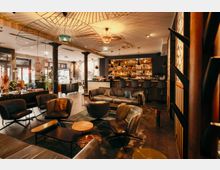 Offener, modern gestalteter Bar- und Loungebereich mit Ledersesseln, Sofas und niedrigen Couchtischen auf Fliesenboden. Im Hintergrund eine lange Theke mit beleuchtetem Flaschenregal, schwarze Säulen und auffällige, fächerförmige Deckenleuchten in warmem Licht.