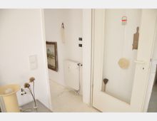 Ein schlichter Innenraum mit weißen Wänden, einem Glasabschnitt in der Tür und einem an der Wand befestigten Heizkörper. An der Wand hängen ein Gemälde, ein kleiner Besen und andere Gegenstände; auf einem Tisch im Vordergrund stehen eine Tischlampe und ein kleines Gefäß mit getrockneten Blumen.