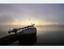 Ein Segelboot liegt an einem Steg auf einem ruhigen See, umgeben von Morgennebel. Im Hintergrund sind Silhouetten von Bäumen und ein heller Sonnenaufgang zu erkennen.