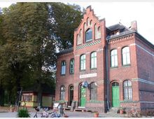 Backsteingebäude eines kleinen historischen Bahnhofs mit grünen Türen und Fenstern, einer überhängenden Bahnhofsuhr und einem Schild mit der Aufschrift „Mellensee-Saalow“. Im Vordergrund stehen Fahrräder, links daneben ein kleiner Kiosk unter großen Bäumen.