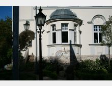 Helle Fassade eines klassischen Gebäudes mit gewölbtem Erkerdach, großen Fenstern und dekorativen Akzenten. Im Vordergrund stehen schmiedeeiserne Laternen und eine Metallbogenstruktur, umgeben von gepflegten Büschen und Bäumen.