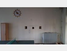 Ein einfacher Innenraum mit einer cremefarbenen, leicht beschädigten Wand. An der Wand befinden sich eine Uhr in Form eines Steuerrads, drei kleine rechteckige Öffnungen und ein Heizkörper unterhalb. Im Vordergrund ist ein Teil einer Tischtennisplatte sichtbar, links steht ein Holzschrank.