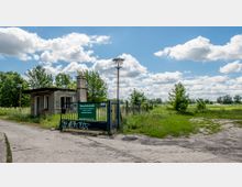 Eingang eines Tierschutzgeländes mit einem kleinen, einfachen Wachhäuschen und einem blauen Metalltor, auf dem ein Schild mit Informationen zum Tierschutzverein angebracht ist. Im Hintergrund Wiese, Bäume und ein bewölkter Himmel.