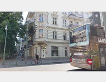Ecke einer belebten Innenstadtstraße mit einem historischen Wohnhaus im Stil des Neoklassizismus, verziert mit Balkonen und Stuckelementen. Im Vordergrund fährt ein Doppeldeckerbus vorbei, daneben ein Radfahrer und mehrere geparkte Fahrräder unter Bäumen.