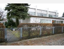 Wohnhaus mit modernistischer Architektur, flachem Dach und großen Fenstern. Vor dem Gebäude befinden sich ein Metallzaun, eine Hecke und ein kleiner Garten mit einem Nadelbaum.