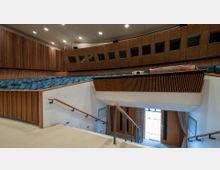 Innenraum eines modernen Hörsaals mit ansteigend angeordneten blauen Sitzreihen und Holzwandverkleidung. Im Vordergrund befindet sich eine Treppe mit Holzgeländern, die zu einer Eingangstür führt; darüber eine Empore mit technischer Ausstattung.