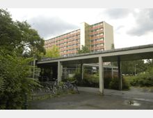 Ein mehrstöckiger Wohnblock in hellen Gelb- und Orangetönen, umgeben von Bäumen und Sträuchern, im Vordergrund ein überdachter Gehweg mit Fahrradständern und mehreren abgestellten Fahrrädern.