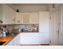 Helle Küche mit weißen Einbauschränken in L-Form, Holzarbeitsplatte und Edelstahlspüle an der linken Seite. An der Rückwand sind ein Fliesenspiegel, eine Abtropfschale und Küchenutensilien zu sehen; rechts steht ein hoher Vorratsschrank neben einer weißen Tür.