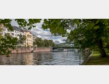 Ein Fluss mit einem Ausflugsboot im Vordergrund, umgeben von grünen Bäumen und einem Park auf der rechten Seite. Links im Bild stehen mehrstöckige Wohnhäuser, und im Hintergrund ist eine grüne Brücke mit Industriekaminen zu erkennen.