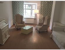 Wohnzimmer mit Holzboden, zwei hellbeigen Ohrensesseln, einem weißen Sofa, einem kleinen cremefarbenen Tisch und einem weißen Sekretär. Im Hintergrund Fenster mit gemusterten Vorhängen und einer Stehlampe.