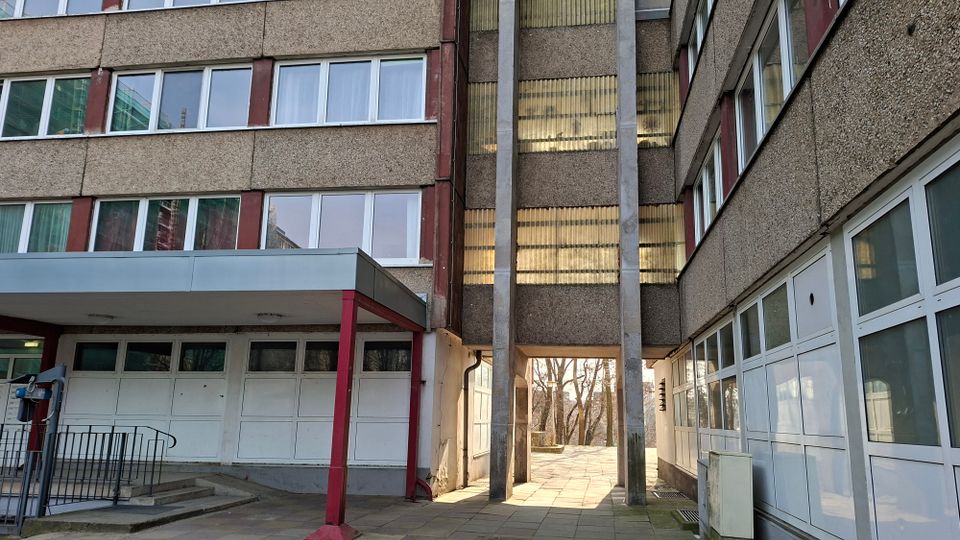 Innenhof zwischen zwei mehrstöckigen Betongebäuden mit langen Fensterbändern und einem überdachten Eingang mit Stützen. In der Mitte führt ein Durchgang unter einem verbindenden Gebäudeteil nach außen, flankiert von hohen Betonpfeilern und gepflastertem Boden.