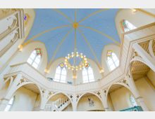 Innenraum einer Kirche mit gewölbter, himmelblauer Decke und goldenen Sternenornamenten. Große Fenster mit buntem Glas und ein zentraler Kronleuchter prägen den Raum, umgeben von Emporen mit filigranen Verzierungen.