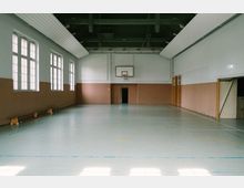 Innenansicht einer leeren Sporthalle mit hellem Boden und beige gestrichenen Wänden. Markierungen für Spielfelder sind auf dem Boden zu sehen, an der Stirnseite befindet sich ein Basketballkorb oberhalb einer Tür. Links befinden sich große Fenster, die Tageslicht hereinlassen.