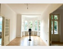 Helles Wohn- oder Arbeitszimmer mit großen Sprossenfenstern und einem Erkerbereich, der viel Tageslicht einlässt. Der Raum hat einen hellen Parkettboden, weiße Wände, Heizkörper unter den Fenstern und eine Glastür zur Seite.