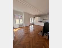 Heller, leerer Wohnraum mit Fischgrätparkettboden, hohen Decken und großen, bogenförmigen Fenstern, die Tageslicht hereinlassen. Im Hintergrund befindet sich eine kleine erhöhte Fläche mit einer Metallleiter und einer weißen Trennwand.