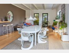 Wohnzimmer mit modernem Design: Ein großer weißer Esstisch mit geschwungenen Stühlen steht auf einem Teppich, umgeben von zahlreichen Zimmerpflanzen. Im Hintergrund befinden sich ein graues Sofa mit bunten Kissen, eine dunkelbraune Wand mit Regal sowie drei Fenster, die Tageslicht hereinlassen.