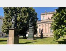 Denkmalgruppe mit drei historischen Statuen auf Sockeln in einem parkähnlichen Bereich, umgeben von Bäumen. Im Hintergrund ist ein klassizistisches, rosafarbenes Gebäude mit Säulen und Statuen auf dem Dach zu sehen.