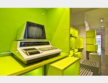 Ausstellungsraum mit einem historischen Computer, dem Commodore PET 2001, in einer gelben Vitrine. Im Hintergrund sind weitere gelbe Vitrinen mit elektronischen Geräten und ein heller Durchgang zu sehen.