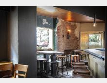 Innenraum eines Cafés mit rustikalem Design, geprägt von unverputzten Backsteinwänden, Holzstühlen und -tischen sowie einer Theke aus Holz. Große Fenster lassen Tageslicht herein, und eine hängende Glühbirne sowie eine Wandlampe sorgen für zusätzliche Beleuchtung.