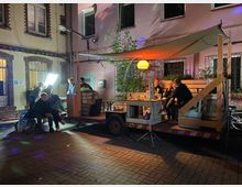 Ein beleuchteter Innenhof mit einer mobilen Holzplattform auf einem Anhänger, auf der einige Personen auf Sitzbänken sitzen und ein runder, orange leuchtender Lampenschirm hängt. Im Hintergrund befinden sich rosa- und gelbfarbene Hauswände mit großen Fenstern und Gittern.