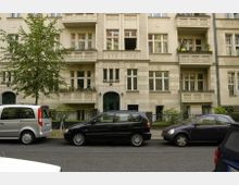 Mehrstöckiges Wohngebäude im historischen Stil mit hellen Fassaden und mehreren Balkonen, davor parkende Autos und ein Baum am Straßenrand.