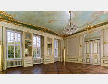 Ein prächtiger Barocksaal mit verzierten Stuckelementen in Gold und pastellfarbigen Wänden. Große Fenster mit Blick nach draußen, ein kunstvoll bemalter Himmel an der Decke und ein Kristalllüster prägen den Raum mit einem Parkettboden.