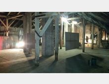 Ein hölzerner Dachboden mit sichtbaren Holzbalken und Holzdielenboden, teilweise beleuchtet. Im Raum sind Kunstwerke und Leinwände zu sehen, sowie eine beleuchtete Treppe im Hintergrund.