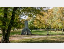 Steinmonument auf einer Grünfläche in einem parkähnlichen Gelände, umgeben von zahlreichen Bäumen mit herbstlichem Laub. Im Vordergrund ist ein Weg sichtbar, der durch den Park führt.