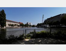 Uferansicht eines Flusses mit geländerbegrenztem Weg im Vordergrund, links ein historisches Gebäude mit rotem Dach, rechts moderne Bürogebäude und ein Kran. Im Hintergrund sind Wohnhäuser und Bäume sichtbar, während Boote am rechten Ufer liegen.