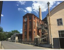 Backstein-Industriegebäude mit hohen Fenstern, einer außenliegenden Metalltreppe und einem hohen Schornstein. Daneben befinden sich eine beige Wand mit Fenstern und ein Schild mit der Aufschrift „Deutsches Leder Museum“ auf einer Brücke über der Straße.