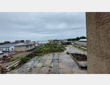 Industrie- oder Gewerbegebiet mit verfallenen Betonflächen, bewachsenen Erdaufschüttungen und mehreren Lagerhallen; im Hintergrund sind Siedlungsgebäude und ein Kirchturm sichtbar. Der Himmel ist bewölkt.