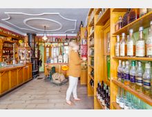 Ein historischer Tante-Emma-Laden mit Holzregalen, die alte Flaschen und Produkte ausstellen. Im Hintergrund stehen ein gusseiserner Ofen und eine Theke, während ein unscharf fotografierter Mensch die Regale betrachtet.