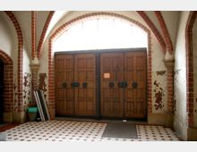 Eingangsbereich eines historischen Gebäudes mit großen, massiv verzierten Holztüren unter einem halbrunden Fenster. Die Wände zeigen teilweise abblätternde Farbe, und die Decke hat rote Backsteinbögen, die auf steinernen Kapitellen ruhen. Der Boden ist mit einem rot-weiß gemusterten Fliesenmuster gestaltet.