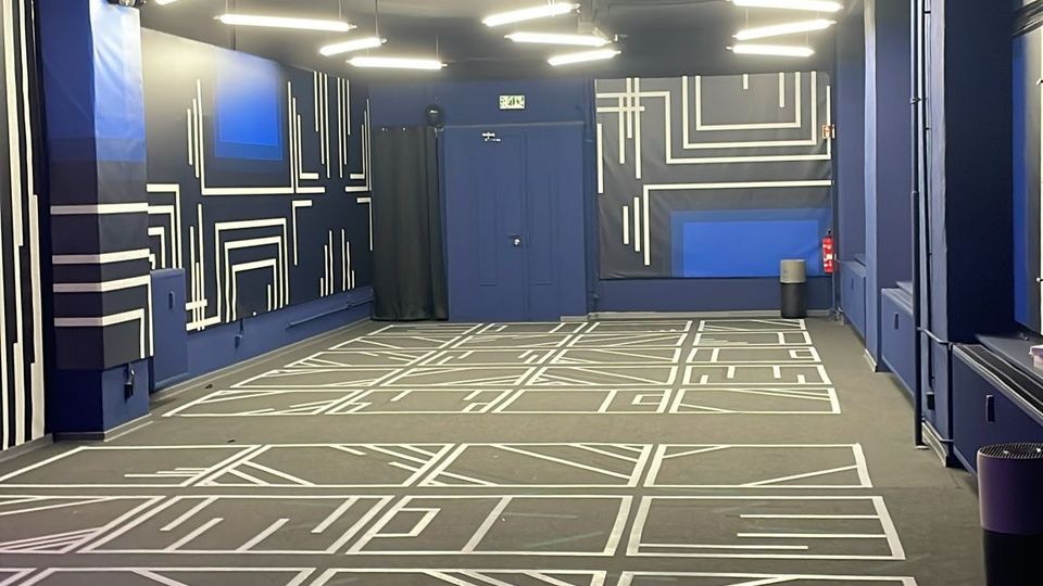 Langer, moderner Innenraum mit dunkelblauen Wänden und grauem Boden, der mit weißen geometrischen Linienmustern gestaltet ist. An der schwarzen Decke hängen mehrere lineare LED-Leuchten; am Ende des Raums befindet sich eine blaue Tür.