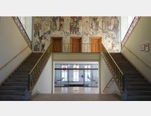 Großzügiger Treppenaufgang in einem Gebäude mit symmetrisch angeordneten Treppen, die zu einem Geländer mit drei Holztüren führen. Über den Türen ist ein großes Wandgemälde mit bunten Figuren dargestellt, und darunter befindet sich ein Durchgang mit Glastüren, durch die ein heller Raum mit Fenstern und einem Tisch sichtbar ist.