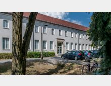 Zweigeschossiges Gebäude mit hellgrauer Fassade, zahlreichen symmetrisch angeordneten weißen Fenstern und einem roten Satteldach. Im Vordergrund stehen parkende Autos, ein Fahrrad sowie Bäume, die teilweise den Blick auf die Fassade verdecken.