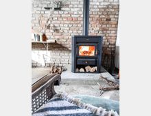 Ein gemütlicher Wohnbereich mit einem schwarzem Kaminofen vor einer Wand aus rohen Ziegelsteinen. Auf der Ofenbank liegen Holzscheite, daneben sind Regale mit Dekorationsgegenständen und Büchern angebracht, und im Vordergrund ist ein gepolsterter Stuhl mit einer gestreiften Decke sichtbar.