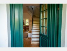 Blick durch eine geöffnete grüne Eingangstür in einen Hausflur mit Holzdielenboden und einer geschwungenen Treppe mit weißen Stufen und Holztrittkanten. Im Hintergrund ist ein heller Wohn- oder Essbereich mit Tisch sowie ein Fenster zu sehen.