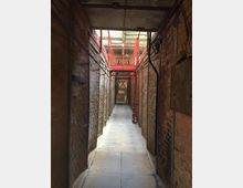 Ein schmaler, gemauerter Gang aus roten Ziegeln mit freiliegenden Kabeln an den Wänden. Der Gang führt zu einer Metallstruktur mit roten Geländern unter einem Dach aus Wellblech.