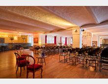 Großer Veranstaltungsraum mit Holzboden, niedriger Decke und weißen Stützpfeilern, bestuhlt mit mehreren Reihen schwarzer Stühle. Links befindet sich eine Bar mit dekorativer Wandgestaltung und Kronleuchtern, im Hintergrund große Fenster mit roten Vorhängen.