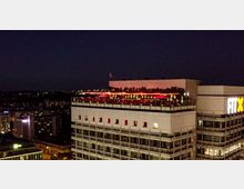 Ein Hochhaus in einer Stadt bei Nacht, dessen Dachterrasse mit roten Lichtern beleuchtet ist und von vielen Menschen besucht wird. Im Vordergrund ist der Schriftzug „FitX“ an einem Nachbargebäude zu sehen, umgeben von weiteren beleuchteten Gebäuden.