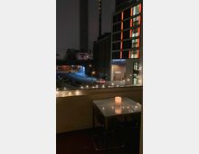 Ein kleiner Balkon bei Nacht mit einem Tisch aus Glas, auf dem eine brennende Kerze steht. Im Hintergrund sind mit Lichtern beleuchtete Gebäude, darunter ein Hotel, sowie eine Straße mit spärlichem Verkehr und einer Bahnbrücke zu sehen. Lichterketten schmücken die Balkonbrüstung.