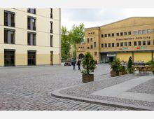 Pflastersteiner Platz mit Blick auf ein gelbes, vierstöckiges Wohn- oder Geschäftsgebäude links und eine historische gelbe Backsteinhalle mit der Aufschrift „Flaschenbier-Abteilung“ rechts. Im Vordergrund befinden sich Blumenbeete, Sitzgelegenheiten und Pflanzkübel mit kleinen Bäumen.