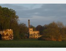Historisches Gebäude mit gelblichem Putz und einem hohen Schornstein, eingebettet in eine bewaldete Parklandschaft mit Wiesen im Vordergrund; links im Bild Reste einer Ruinenmauer.