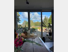 Blick aus einem modernen Wohnraum durch bodentiefe Glasfenster auf einen Garten mit Pflanzen, Bäumen und einem gepflasterten Weg. Im Vordergrund stehen ein Holztisch mit Dekorationen und eine Vase mit bunten Kunstblumen.