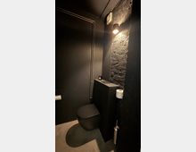 Kleines, dunkel gestaltetes Gäste-WC mit schwarzer Wandtoilette, Betonboden und sichtbaren Metallrohren an der Wand. Eine freigelegte Ziegelwand mit Wandleuchte, schmalem Ablagekasten sowie Toilettenpapierhalter und WC-Bürste ergänzen den Raum.