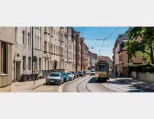 Straßenszene mit einer Straßenbahn, die in Richtung „Hauptbahnhof“ fährt, umgeben von mehrstöckigen Wohnhäusern im traditionellen Baustil. Am Straßenrand parken Autos, während Gehwege und vereinzelte Bäume die Stadtkulisse ergänzen.
