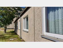 Außenansicht eines einfachen Wohngebäudes mit rauer Putzfassade, mehreren weißen Fenstern mit Spitzenvorhängen und einem Fallrohr an der Wand. Im Vordergrund ist ein kleiner Rasen mit einem Baum sichtbar.